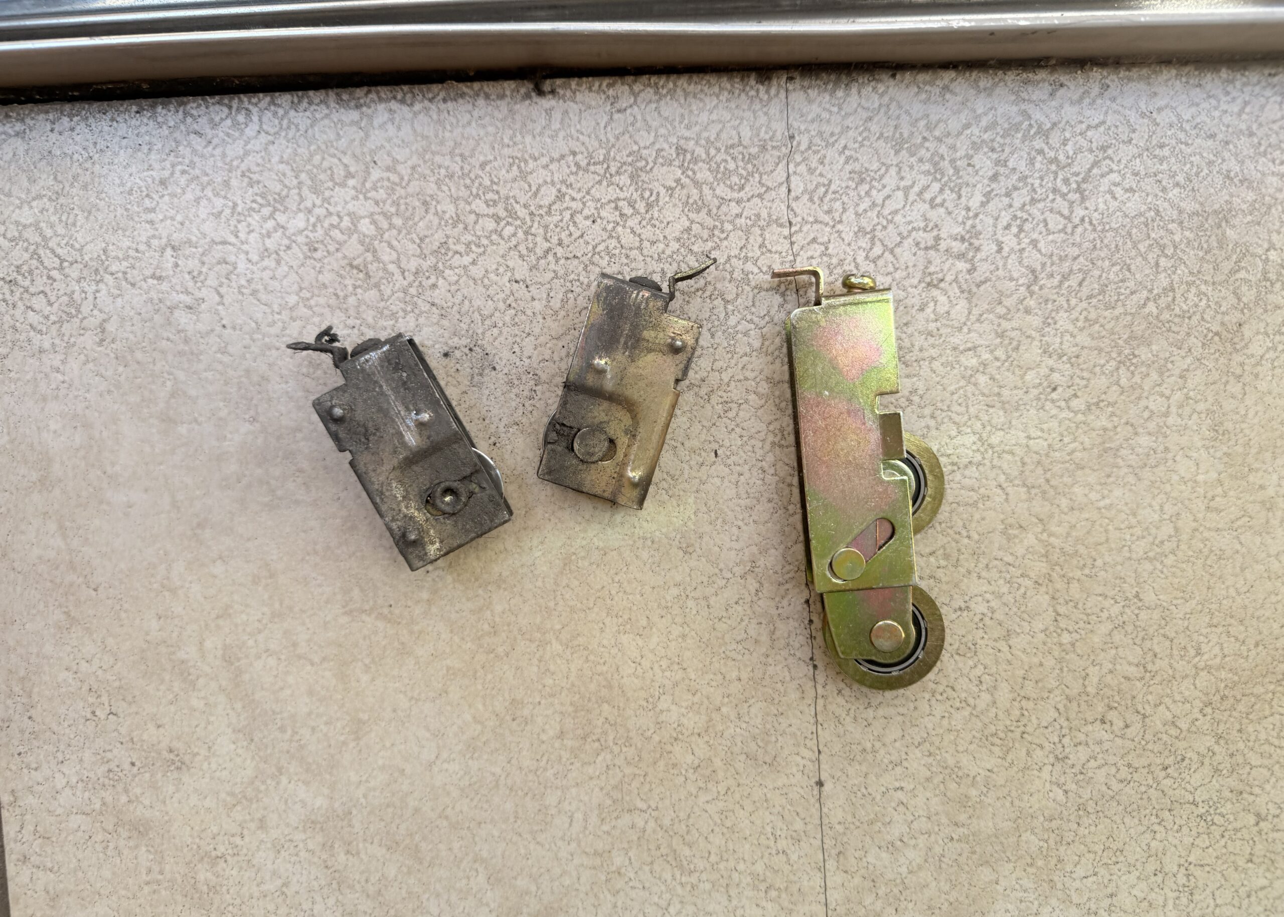 Three metal sliding door parts, two old and corroded on the left, and one newer, cleaner part with two wheels on the right, all placed on a light-colored textured surface.