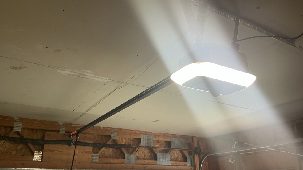 Interior view of a garage with a newly installed garage door opener mounted to the ceiling, showing the opener light turned on and the rail system connected above the door.