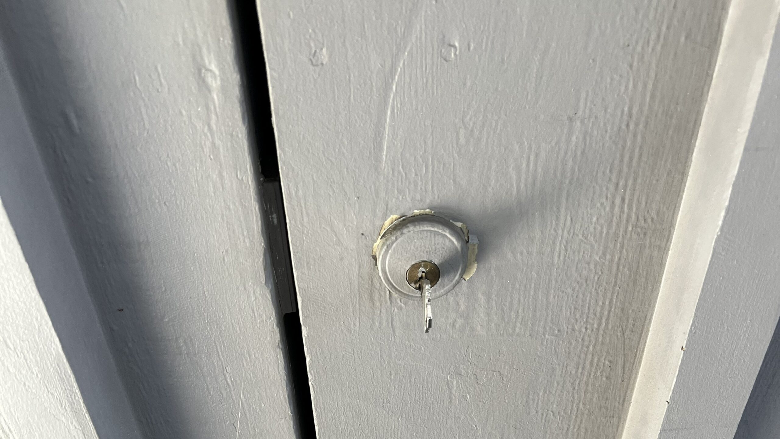 Close-up of a weathered white wooden door with a small round metal lock, narrow dark gap, and stained concrete threshold below.