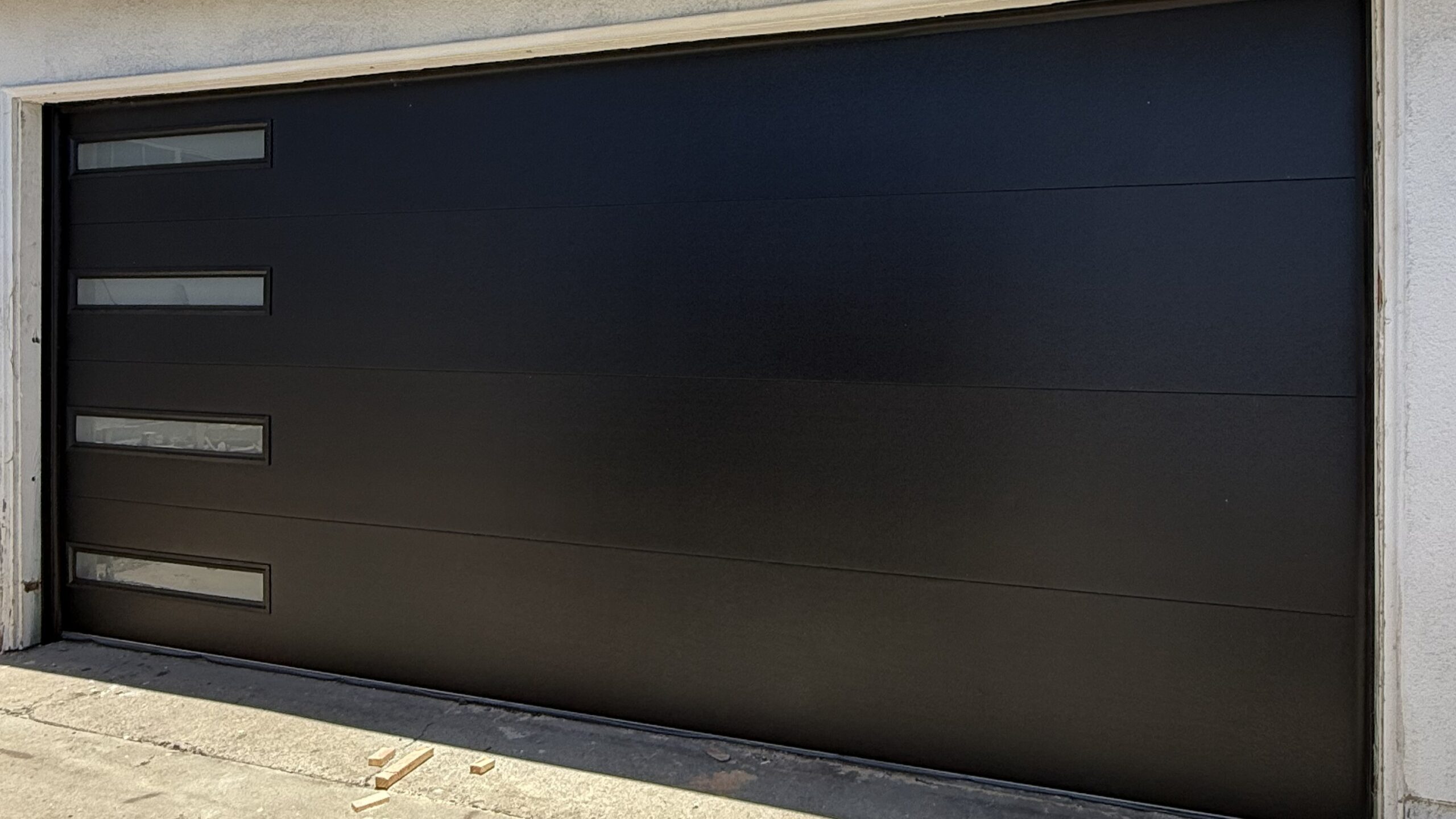 Closed dark charcoal sectional garage door set in a concrete facade, with three narrow frosted windows on the left, lit by bright daylight.